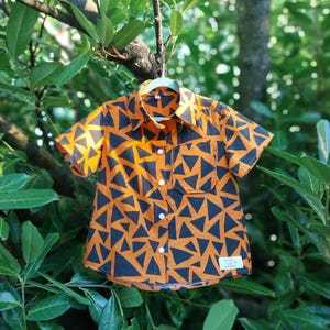 May include: Short-sleeved button-down shirt with an orange background and a repeating pattern of dark blue triangles. The shirt has a collar, a chest pocket, and white buttons. The shirt is hanging on a hanger.