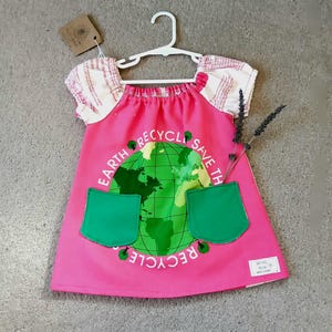 May include: A pink dress with green pockets and the words "Earth Recycle Save The" encircling a green globe. The dress has white and pink patterned sleeves and is hanging on a white hanger.
