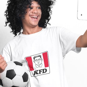 May include: A person wearing a white t-shirt with a red and white logo that says "KFD" and a cartoon image. The person is holding a soccer ball and a tablet. The person has a black curly wig.