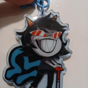 May include: A clear acrylic keychain featuring a cartoon character with black hair, red-tinted sunglasses, and a wide grin. The character is dressed in black and red, holding a red and silver object. A blue emblem is on the left side of the keychain.