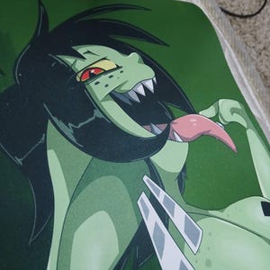 May include: A green mousepad featuring a cartoon character with green skin, black hair, and a long pink tongue. The character has sharp teeth and red eyes. The background is a solid green color.