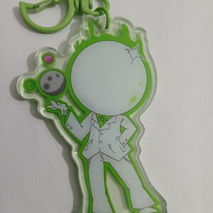 May include: A clear acrylic keychain featuring a cartoon figure in a white suit with a green outline. The figure has a blank white face and is holding a moon-shaped object. The keychain has a green clasp.