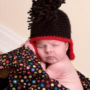May include: A baby wearing a black and red crocheted hat with a mohawk style. The hat has a red trim and a black pom-pom on top. The baby is lying on a black and white polka dot blanket.