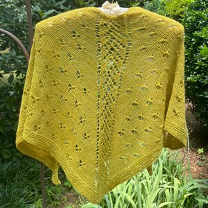 May include: A yellow knitted shawl with a lacy pattern. The shawl is hanging on a hanger and has a triangular shape.