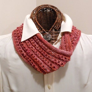 May include: A pink and orange knitted infinity scarf with a delicate stitch pattern. The scarf is displayed on a white shirt.