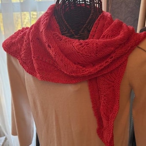 May include: A vibrant red knitted shawl draped over a mannequin. The shawl features a delicate openwork pattern along the edges and a textured knit throughout. The bright red color contrasts with the neutral-toned mannequin and background, highlighting the shawl's intricate design.