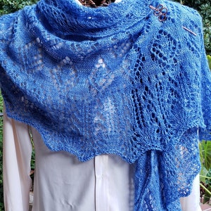 Puede incluir: Un chal de punto azul con un delicado patrón de encaje. El chal está drapeado sobre una camisa blanca y sujeto con un broche decorativo.