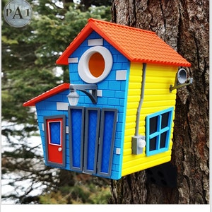 May include: A colourful birdhouse shaped like a small house with a blue, yellow and red exterior. The birdhouse has a red roof, a round entrance hole and a small window.