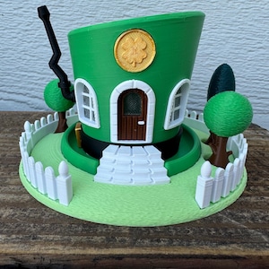 May include: A whimsical green hat-shaped house with a white picket fence, trees, and a shamrock emblem. The house features a brown door, white windows, and a black chimney. The base is light green.
