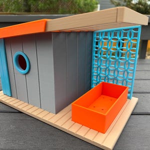 May include: A contemporary birdhouse with a grey body, orange roof trim, and a light blue accent. It has a circular opening and a decorative blue lattice panel. An orange rectangular box sits on the wooden base.
