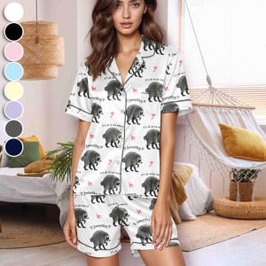 Op de afbeelding: Een witte pyjamaset met korte mouwen en knoopsluiting, voorzien van een herhalend patroon van grijze wasberen, kleine rode hartjes en de tekst "it's ok to be weird" en "Jimothy" in zwarte letters met donkergrijze biesafwerking.