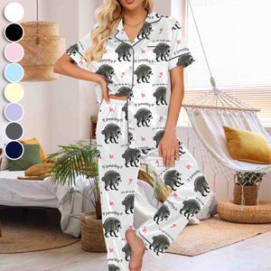 Op de afbeelding: Een witte pyjamaset met een herhalend patroon van grijze wasberen, kleine rode hartjes en de tekst "Jimothy" en "it's ok to be weird". De top is een overhemd met korte mouwen en een kraag, en de broek is een bijpassende lange broek.