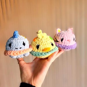 Peut inclure: Trois jouets en peluche en crochet en forme de planètes. Le premier est bleu clair avec un anneau rose, le deuxième est jaune avec un anneau vert et le troisième est rose avec un anneau violet.
