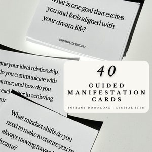 May include: A set of 40 guided manifestation cards with black text on white cards. The cards are stacked on a white surface. The text on the top card reads "What is one goal that excites you and feels aligned with your dream life?"