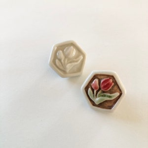 以下が含まれることがあります： チューリップのレリーフ模様が施された、セラミック製の六角形のタイル2枚。1枚は白地に白のチューリップ、もう1枚は茶色地にピンクのチューリップです。