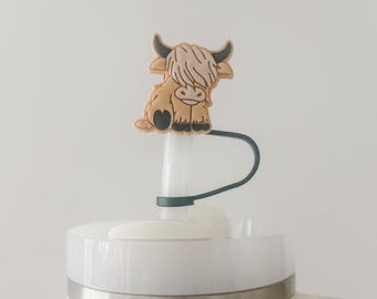 May include: White Stanley travel mug with a black and green lid and a brown highland cow straw topper. The mug has a silver band around the top and the Stanley logo is visible on the front.