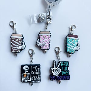 May include: Five badge reels featuring colorful silicone charms. Three charms depict skeleton hands holding coffee cups in brown, pink, and teal. Two square charms feature skeleton graphics with the text I CAN'T LIVE LAUGH LOVE IN THESE CONDITIONS and HAVE THE DAY YOU DESERVE.