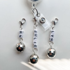 May include: Three silver retractable badge reels with silver chains and silver disco ball charms. Each chain has white letter beads spelling out the names "Julie", "Lynn", and "RN".