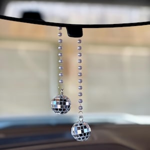 May include: Two small disco ball car charms hanging from a car's rearview mirror. The disco balls are made of mirrored squares and are suspended by strings of silver beads. The background is blurred, suggesting the interior of a vehicle.