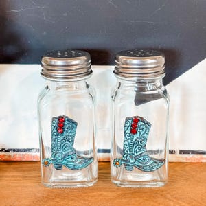 May include: Two clear glass salt and pepper shakers with silver lids. Each shaker features a turquoise cowboy boot design with red and orange accents. The shakers are displayed on a wooden surface.
