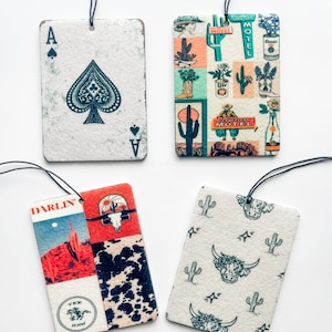 May include: Four different car air fresheners with various designs. The first is a white card with a black ace of spades. The second is a colorful design with cacti, boots, and a motel sign. The third is a collage of red, white, and black with a skull, cacti, and the words "Darlin'" and "Yee Haw". The fourth is a white card with a green cactus and a cow's head pattern.