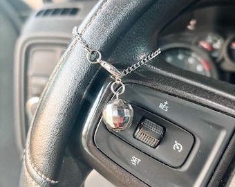 May include: A silver chain with a small, reflective disco ball charm attached to a black steering wheel. The steering wheel has buttons labeled "RES" and "SET". The image is taken in a car.