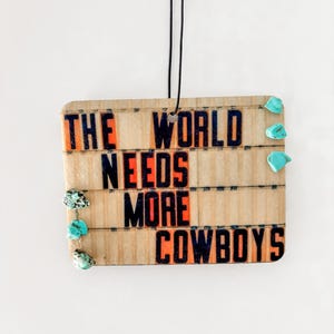 May include: Wooden car air freshener with the text "The World Needs More Cowboys" in black and orange letters. The air freshener is decorated with turquoise stones.