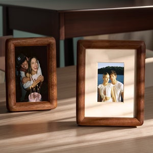 May include: Two wooden picture frames with photos of couples. The frame on the left shows a couple hugging with a birthday cake. The frame on the right shows a couple standing on a jetty by a lake.