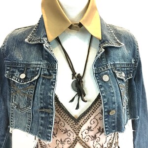 May include: A denim jacket with a brown leather necklace and a beige collared shirt underneath. The jacket has a distressed look and is cropped.
