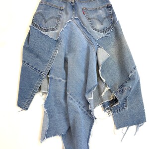 May include: A denim skirt made from repurposed jeans. The skirt is asymmetrical and has a raw edge finish. The skirt is blue and has a brown leather belt.