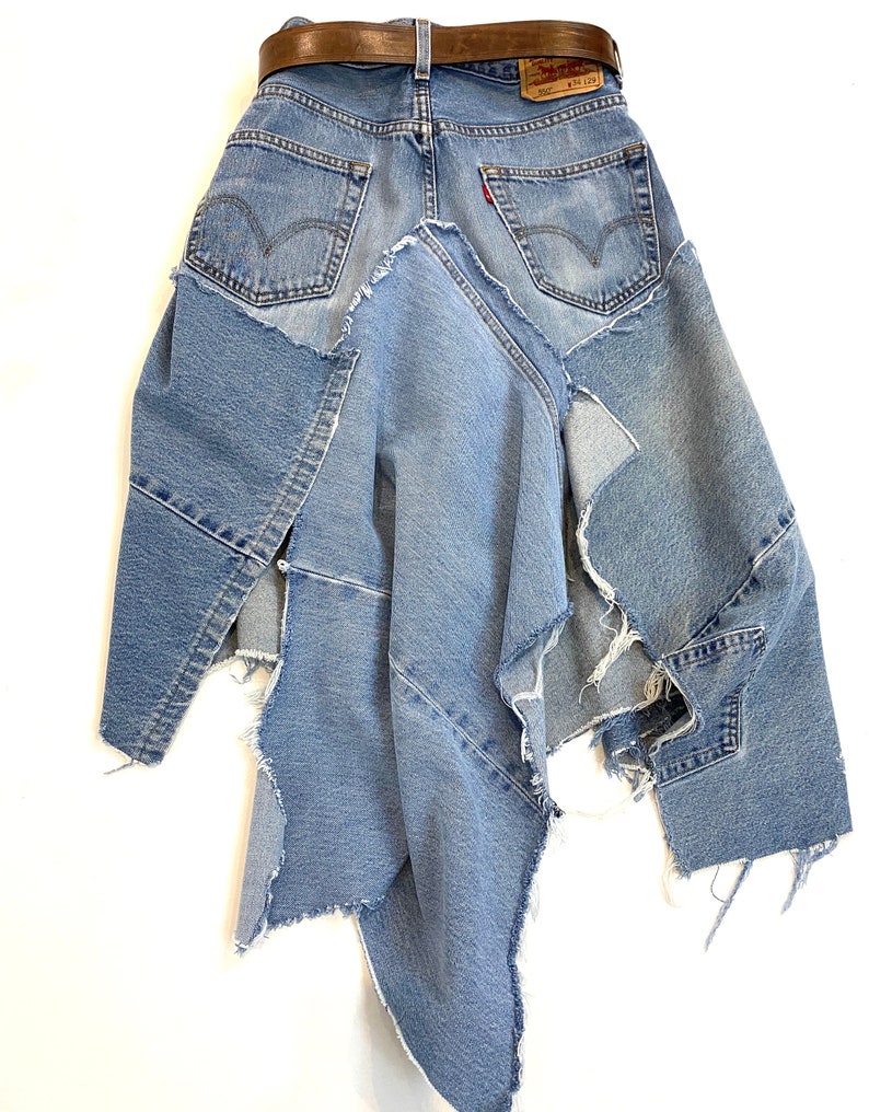 May include: A denim skirt made from repurposed jeans. The skirt is asymmetrical and has a raw edge finish. The skirt is blue and has a brown leather belt.