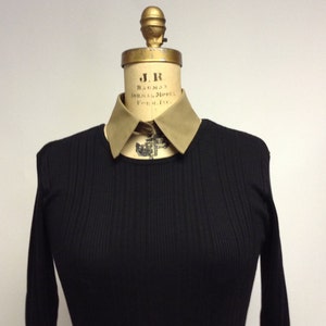 May include: A black ribbed sweater with a tan collar. The collar has the text "J.R. BAUMAN NORMAL MODEL FORMING" on it.