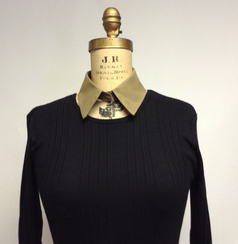 May include: A black ribbed sweater with a tan collar. The collar has the text "J.R. BAUMAN NORMAL MODEL FORMING" on it.