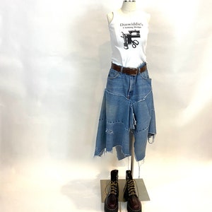 May include: A white tank top with a black graphic of a sewing machine and scissors. The text "Dinwiddie's Clothing Design" is printed on the top. A blue denim skirt with distressed details and a brown belt is worn over the top. The skirt is asymmetrical and has a frayed hem. The model is wearing brown leather boots with laces.