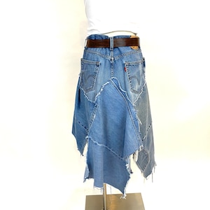 May include: A denim skirt made from repurposed jeans. The skirt is a patchwork of different denim fabrics, with a frayed hem and a brown leather belt.