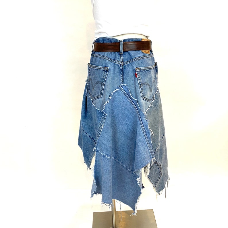 May include: A denim skirt made from repurposed jeans. The skirt is a patchwork of different denim fabrics, with a frayed hem and a brown leather belt.