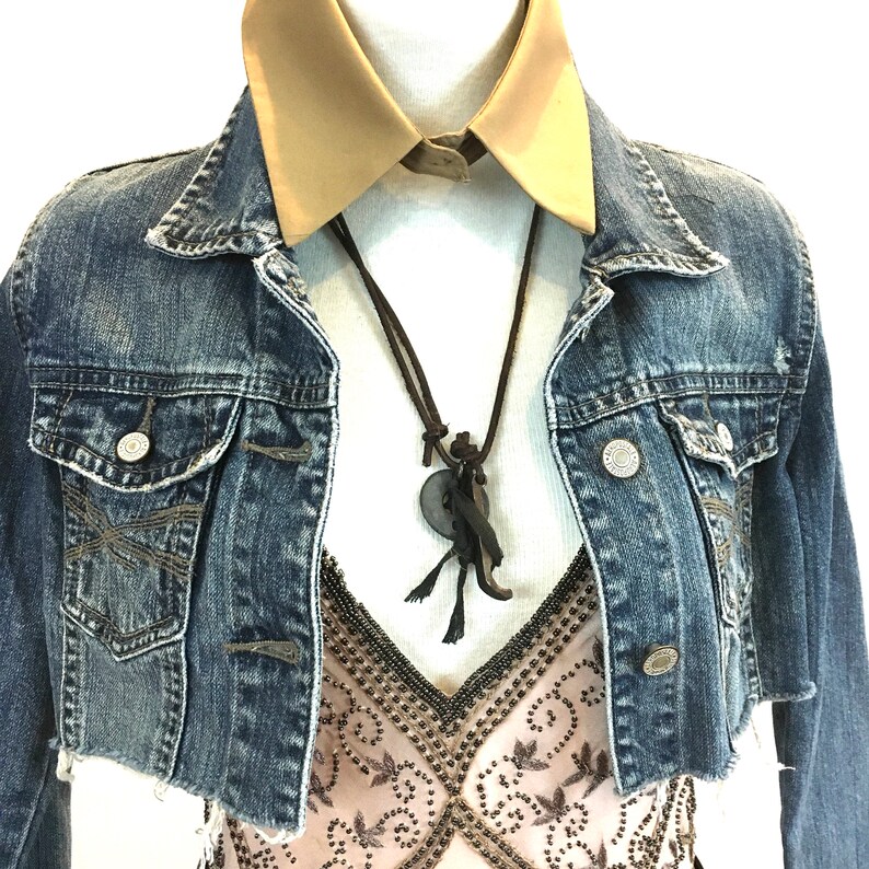 May include: A denim jacket with a brown leather necklace and a beige collared shirt underneath. The jacket has a distressed look and is cropped.