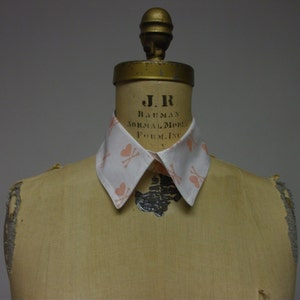 May include: A white collared shirt with a pink and white pattern of hearts and crossbones. The shirt is on a dress form with the text "J.R. BAUMAN NORMAL MODEL FORM, INC. NY" printed on the form.