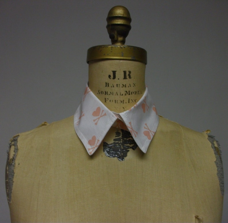 May include: A white collared shirt with a pink and white pattern of hearts and crossbones. The shirt is on a dress form with the text "J.R. BAUMAN NORMAL MODEL FORM, INC. NY" printed on the form.