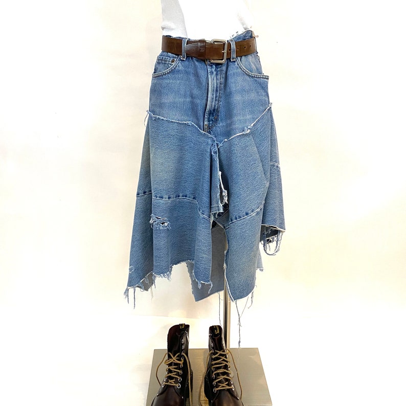 May include: A denim skirt with a brown leather belt and frayed edges. The skirt is made of multiple pieces of denim sewn together. The skirt is displayed on a mannequin with brown leather boots.