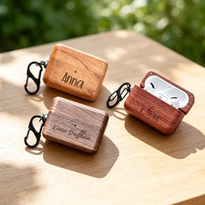May include: Three rectangular wooden cases for wireless earbuds, each featuring a black metal carabiner. The cases are engraved with the names Anna, Kiara Dreffkern, and Olivia. One case is open, revealing white wireless earbuds inside.