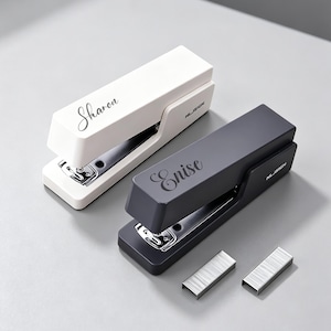 May include: Two desktop staplers, one white and one dark gray, each featuring a custom name in cursive script on the top surface. The white stapler is labeled Sharon and the dark gray one is labeled Enise. Both units include the brand name NUSIGN.