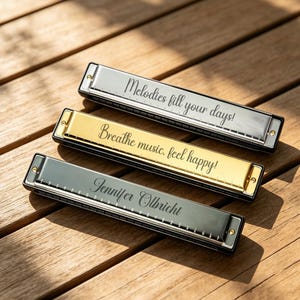 May include: Three harmonicas in silver, gold, and black finishes. The silver harmonica is engraved with "Melodies fill your days!", the gold one with "Breathe music, feel happy!", and the black one with the name "Jennifer Olbricht" in script.