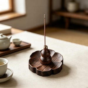 Può includere: Un porta incenso in legno marrone scuro con una base a forma di fiore e un componente centrale a forma di zucca. Un sottile bastoncino di incenso è inserito nell'attacco metallico color ottone sulla sommità della zucca, in posizione verticale.