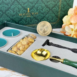 Könnte beinhalten: Ein dunkelgrünes Siegellack-Set mit floralem Design. Das Set enthält teal Kerzen, eine Glasflasche mit Wachsperlen, einen goldenen Wachsstempel mit dem Buchstaben "A" und einen goldenen Löffel mit grünem Griff.