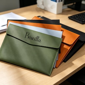 Può includere: Una pila di quattro cartelle rettangolari in materiale liscio. La cartella superiore è verde oliva con il nome Priscilla stampato in corsivo nero. Sotto ci sono cartelle grigio chiaro, arancione brillante e nere, tutte dotate di chiusura a patta.