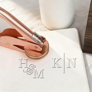 Può includere: Un timbro sigillo in oro rosa con un motivo floreale, premuto su una superficie bianca, creando iniziali in rilievo. Le iniziali "H&M" sono visibili, insieme a "K|N". Un vaso di ceramica bianco è sullo sfondo.
