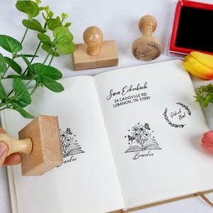 Peut inclure: Plusieurs tampons en bois avec des motifs floraux et une couronne de laurier sont présentés sur une page de carnet. Le texte "Sara Eckenbach" et une adresse sont imprimés sur la page. Un tampon encreur rouge est également visible.