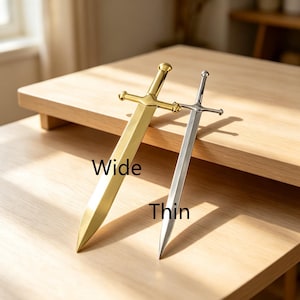 May include: Two metal letter openers shaped like swords. One is gold-toned with a wider blade labeled Wide, and the other is silver-toned with a narrower blade labeled Thin. Both feature a crossguard, a straight handle, and a rounded pommel.