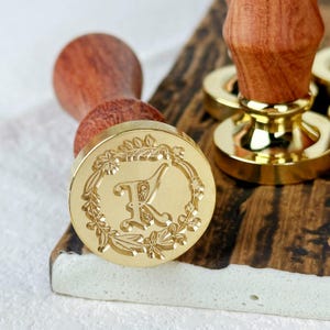 Könnte beinhalten: Ein goldfarbener Wachssiegel-Stempel mit einem floralen Design und dem Buchstaben "K" in der Mitte. Der Stempel ist an einem Holzgriff befestigt.